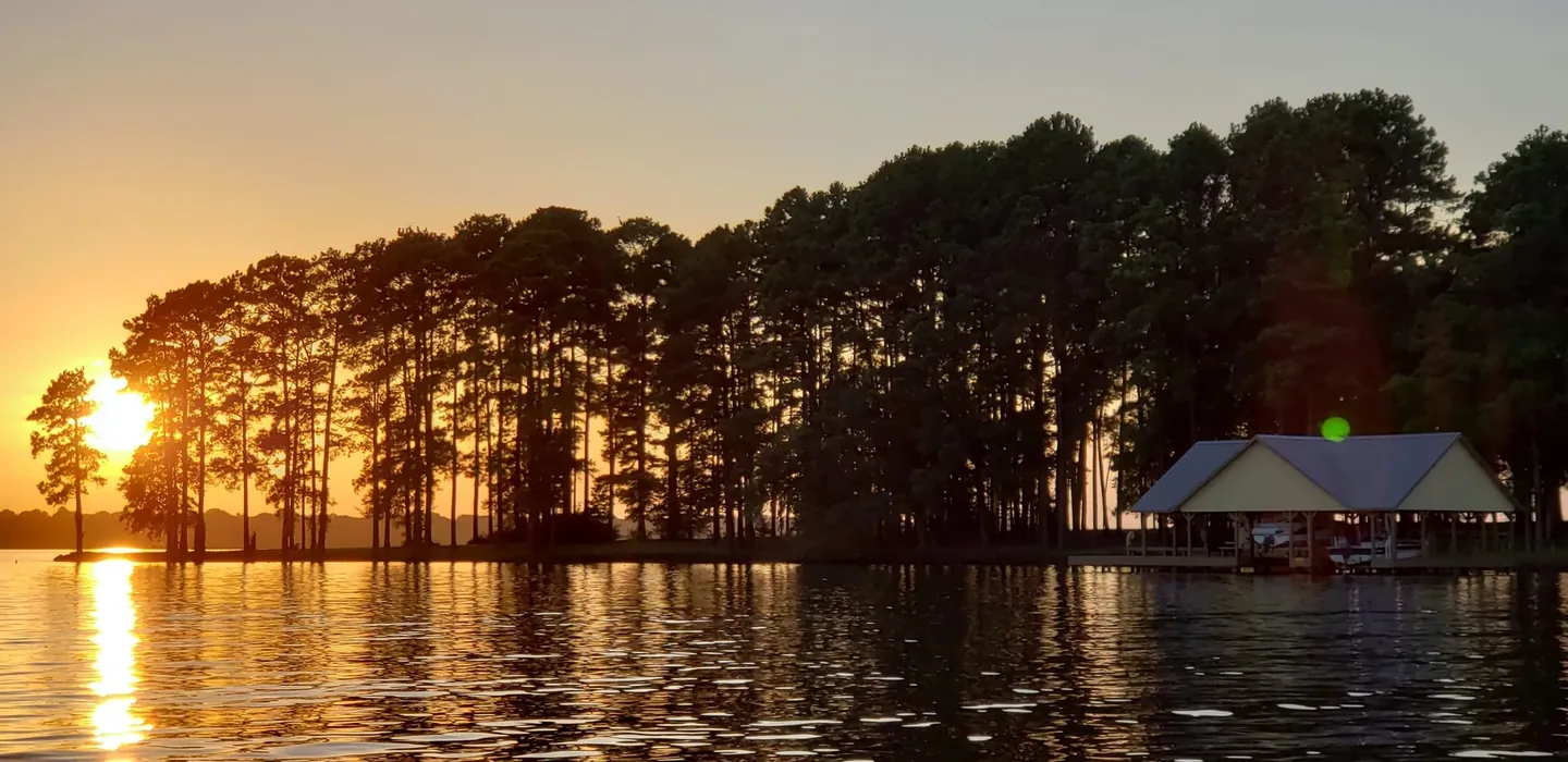 A covered boathouse at the end of a pine-lined point on Lake Palestine at sunset, with calm open water in front of it