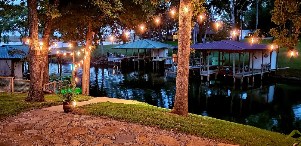String lights over a stone patio at dusk, looking out at covered boathouses on a Lake Palestine cove.